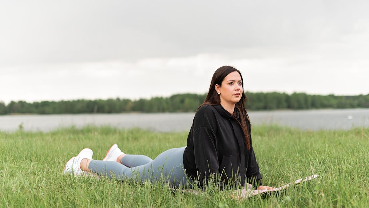 Woman lying on a yoga mat in a grassy field by a lake, promoting relaxation and wellness.